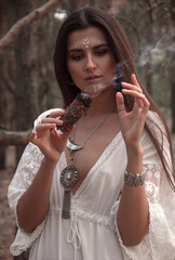 close up young brunette girl in white boho dress and jewelry stands like modern witch with smoky burning stick in hand and makes shaman ritual on forest background. witch cosplay. lifestyle concept