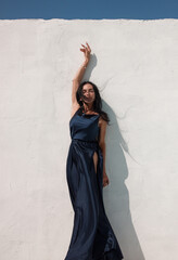 brunette girl in blue long silk dress is standing near white wall background with blue sky at sunny day and smiling cute with hand up in dance. event mood. greek villa. travel concept, free space