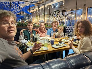 Family of five and baby is sitting at a table in a restaurant. The man in the front is wearing glasses