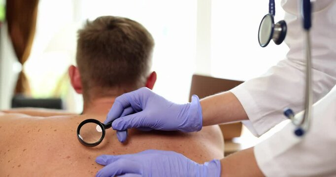 Female professional wearing blue gloves carefully examines patient back using magnifying glass. Skin abnormalities during dermatological screening
