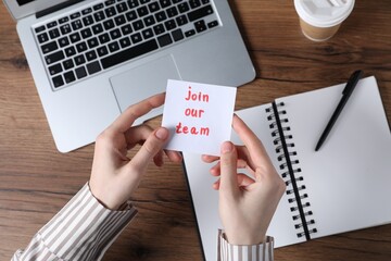Woman holding paper note with words Join Our Team at wooden table, top view