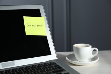 Sticky note with words Join Our Team, laptop and cup on white wooden table against grey background,...