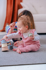 Vertical portrait shows a girl in pink overalls on a grey rug, stacking pastel cups with curiosity...