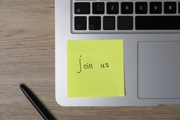 Sticky note with phrase Join Us, laptop and marker on wooden table, flat lay