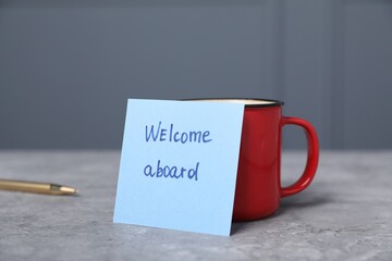 Sticky note with phrase Welcome Aboard, coffee and pen on grey table, closeup