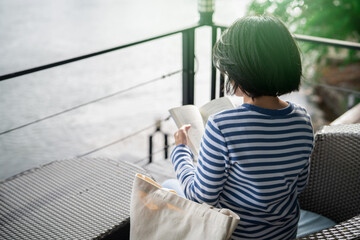 Woman sitting on chair reading a book on balcony overlooking river during golden hour morning. Self...