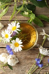 Aromatic herbal tea in glass cup, flowers and green leaves on wooden table, flat lay