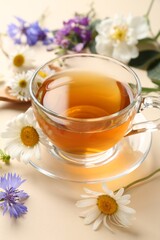 Aromatic herbal tea in glass cup and different flowers on beige background, closeup