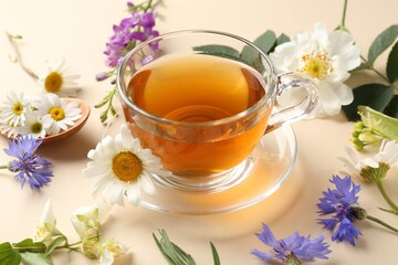Aromatic herbal tea in glass cup and different flowers on beige background, closeup
