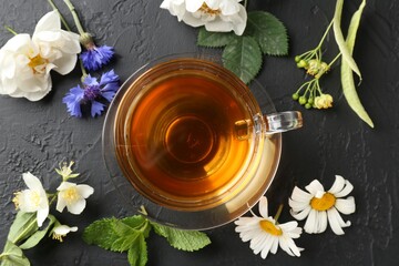 Aromatic herbal tea in glass cup, different flowers and mint on black table, flat lay