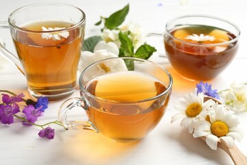 Aromatic herbal tea in glass cups and different flowers on white wooden table, closeup