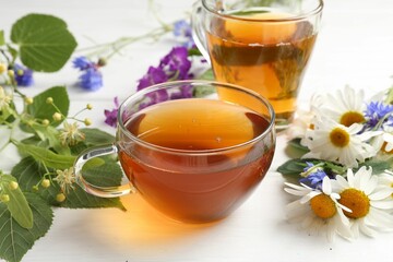 Aromatic herbal tea in glass cups, different flowers and green leaves on white wooden table, closeup