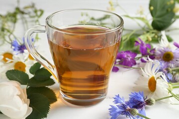 Aromatic herbal tea in glass cup, different flowers and green leaves on white wooden table, closeup