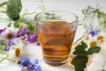 Aromatic herbal tea in glass cup, different flowers and green leaves on white wooden table, closeup