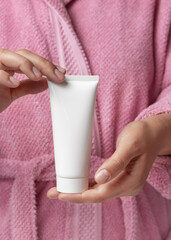 Closeup of woman in pink bathrobe holding white squeeze cream tube in hands, cosmetic mockup