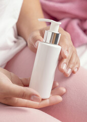 Closeup of female in pink pyjamas holding white cosmetic one pump bottle in hands, lotion mockup