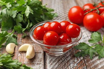 Tasty pickled tomatoes in glass bowl, parsley and garlic cloves on wooden table, closeup