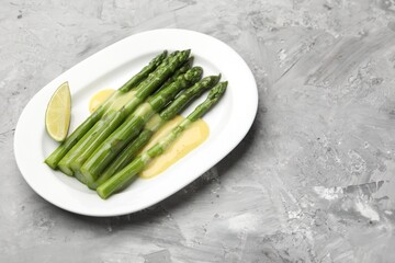 Tasty cooked asparagus served with sauce and lime on grey textured table, space for text
