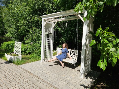 Woman is sitting on a swing in a garden. The swing is white and has a white roof