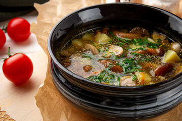 Traditional mushroom soup with potatoes, fresh greens and spices served in a black bowl on parchment paper. Rustic style with cherry tomatoes on the background. Warm and hearty comfort food for menu