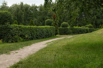 A well-maintained dirt path winds through lush green hedges and open grass in a quiet park. The sky...