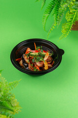 Freshly prepared Asian-style vegetable salad with herbs, served in a black plastic bowl on green background. Colorful and healthy takeaway lunch option