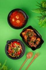 Top view of a complete takeaway lunch with tomato soup and meatballs, vegetable salad, grilled chicken wings and potato wedges in black containers
