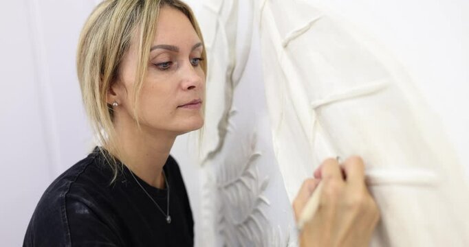 Concentrated woman adds texture by scoring surface of bas relief with small modeling knife. Skilled female carves out main veins of leaves slow motion