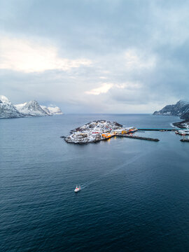 Hus&oslash;y auf der Insel Senja in Norwegen an einem klaren Wintertag mit blauen Farbt&ouml;nen, eisiger Lichtstimmung und einem Fischerboot, das die Insel in Richtung offener See verl&auml;sst