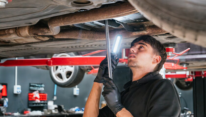 Young male mechanic inspecting vehicle undercarriage with flashlight in auto repair shop