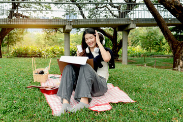 Happy two beautiful Asian women have a picnic in park. Smiling close friends reading a book, drawing a picture outdoor during picnic in green garden. People spend time together in natural surroundings