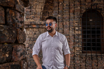 Bearded Latino man posing with attitude in a colonial castle structure in daylight. Fashion, modeling, tourism, and style concepts.