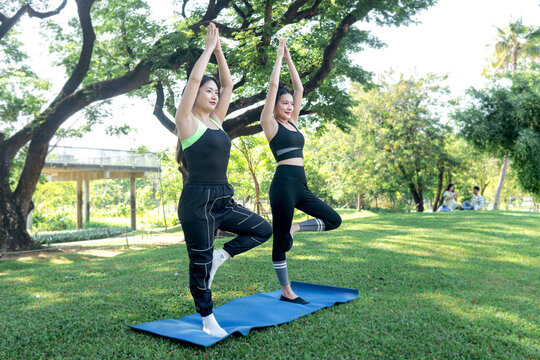 Two Asian women in sportswear stretching body outdoor on yoga mat in park together. Sporty buddy women work out in green garden. Sporty girl engage in yoga healthy exercise with partner in nature.