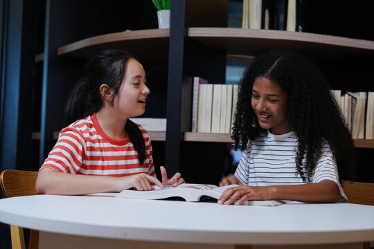 Two teenage girls happily chatting together about their homework and reading books in the school library.
