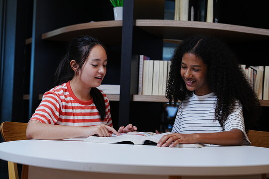 Two teenage girls happily chatting together about their homework and reading books in the school library.