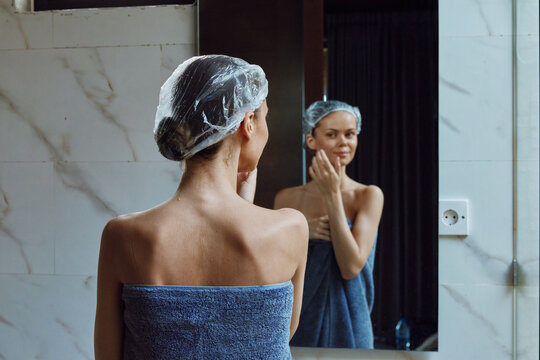 Woman with shower cap wrapped in blue towel touching face in bathroom mirror, skincare routine, personal care, wellness moment, morning self-care.
