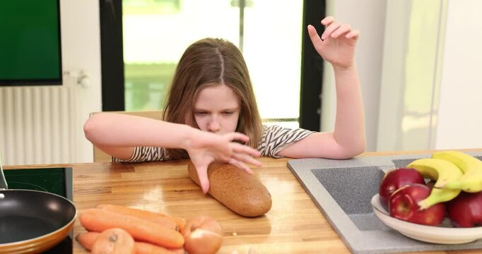 Focused preteen girl plays with bread loaf sitting at wooden table. Bored schoolgirl rotates rye bun near sink waiting for food in domestic kitchen