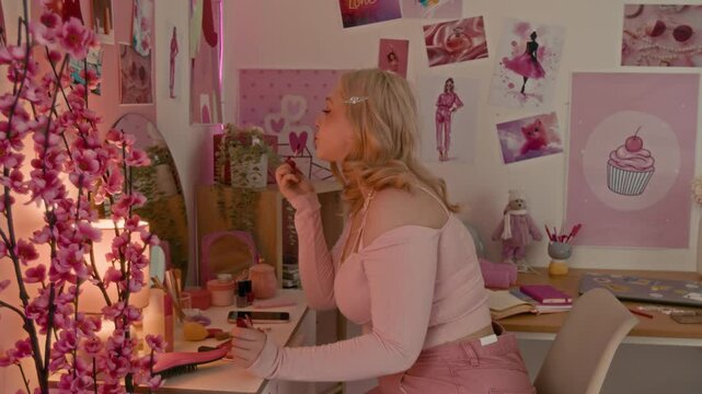Blonde teenage girl applying lipstick while sitting at vanity table in her pink-themed room with numerous posters on walls