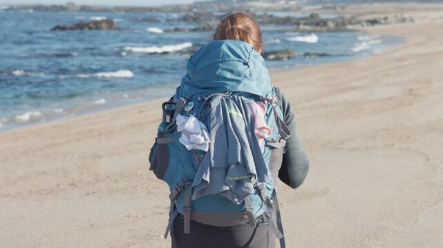Portrait of beautyful midle age woman walking backpack alone by sandy wide long beach and enjoying Atlantic Ocean waves in Portugal. Shot during the Camino Portuguese popular pilgrimage route 4K video