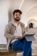 A Latino man wearing orange glasses and curly hair working remotely on a tablet in a castle in a...