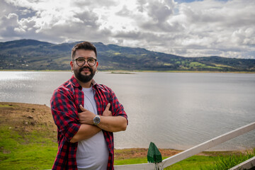 Latino man wearing oversized glasses posing from a viewing platform with Lake Guatavita in the...