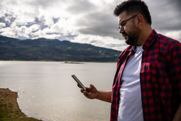 Hipster Latino man in a red striped shirt using his phone by Lake Guatavita. Concepts of...
