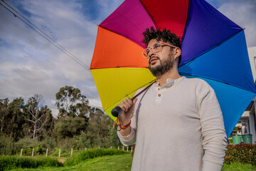 A curly-haired Latino man with orange glasses using a rainbow umbrella showing his support for the...