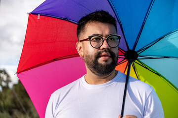 A hipster-style Latino man wearing glasses and holding a rainbow umbrella shows his support for the...