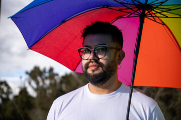 A hipster-style Latino man wearing glasses and holding a rainbow umbrella shows his support for the...
