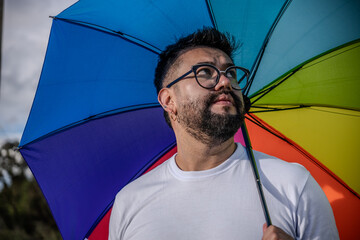A hipster-style Latino man wearing glasses and holding a rainbow umbrella shows his support for the LGBT+ community at the lake in a colonial town in Colombia. Concepts of pride, LGBT, diversity, cele