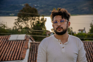Handsome, curly-haired Latino man wearing orange glasses posing by the lake in a colonial town in...