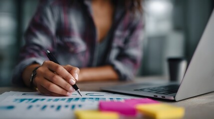 Fototapeta premium Person calculating ROI beside laptop with market news and sticky notes