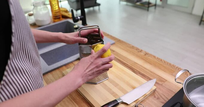 Woman in striped apron grates lemon over wooden cutting board in well-lit kitchen. Large knife and pot on hob indicate active food preparation