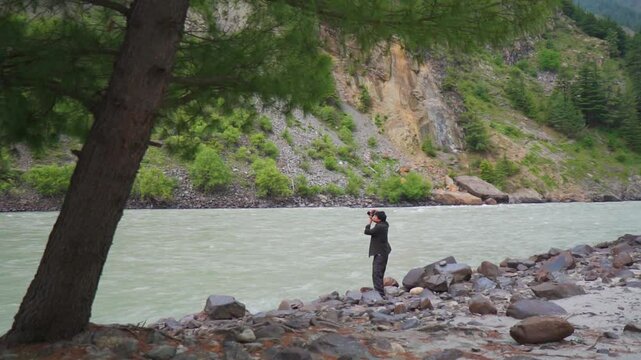 Defocused Indian man taking photo of the Himalayan mountains while standing besides river Chenab near Udaipur in Pattan valley, Himachal Pradesh, India. Tourist take photo of the surroundings in India
